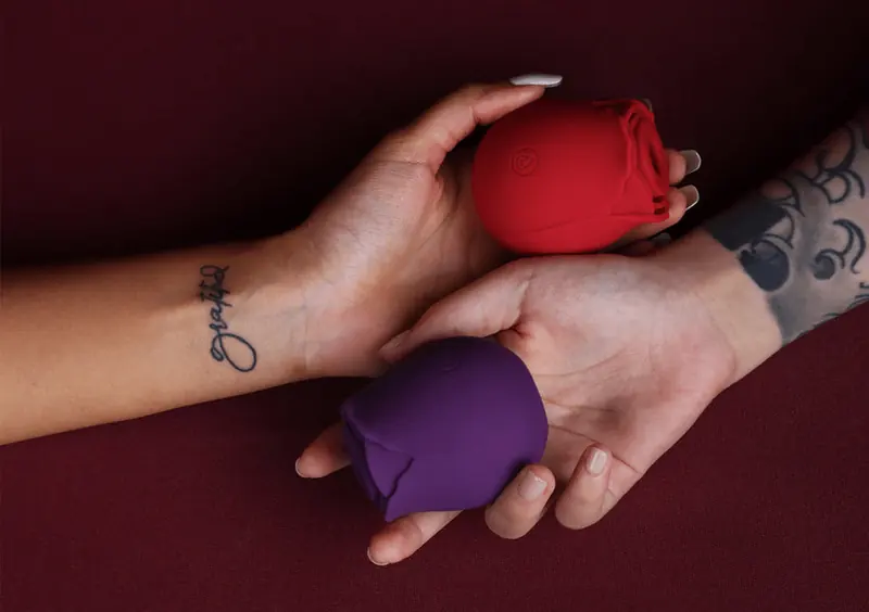 Two adult hands holding red and purple rose-shaped devices over burgundy fabric